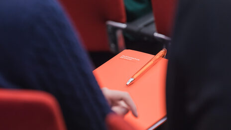 Photo of a person sits on a chair at an event with FH documents on their lap. __ Person sits on a chair at an event with FH documents on their lap.