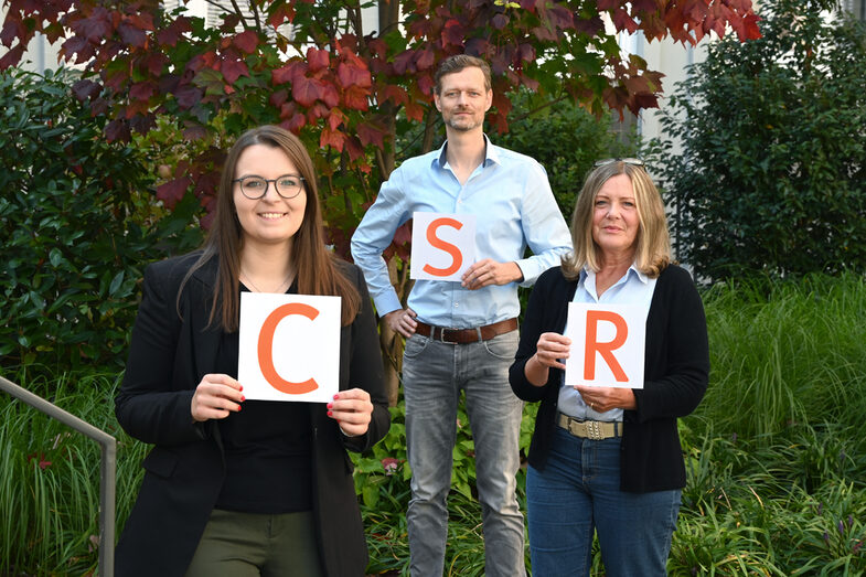 Gruppenbild CSR-Office-Teams: Drei Personen halten jeweils ein Blatt Papier mit je einem orangefarbenen Buchstaben hoch. Von links nach rechts liest man so CSR.