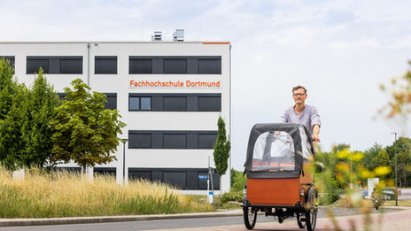 Photo of a man on a cargo bike riding away from a building on the cycle path. The words "Fachhochschule Dortmund" can be seen on the building.