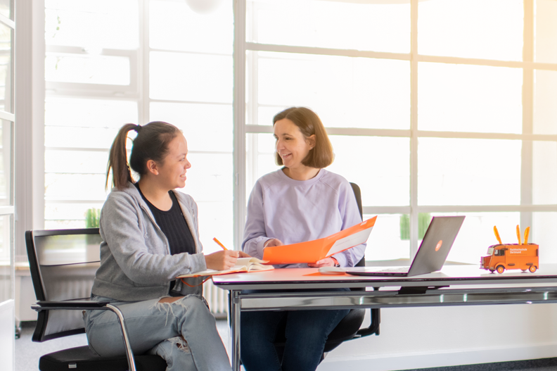 Foto einer Mitarbeiterin der Studienberatung, die eine Studierende berät, die am Tisch neben Ihr sitzt. Sie zeigt dabei auf die Mappe, die Sie in der Hand hält. Die Studierende sieht die Beraterin lächelnd an und macht sich Notizen.