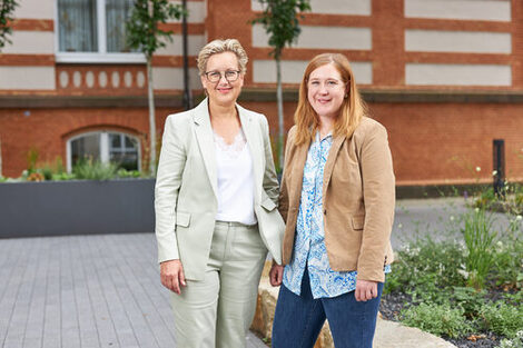 Two women in blazers stand in front of a building and smile for the camera __Two women in blazers stand in front of a building and smile for the camera