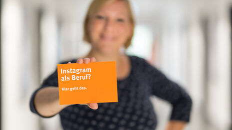 Photo of a woman - blurred - holding an orange postcard from the university of applied sciences up to the camera. The postcard reads "Instagram as a profession? Sure you can.".