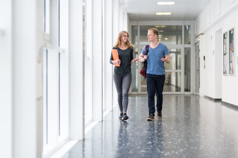 Photo of a female and a male student walking side by side down the hallway and talking lively __One female and one male student walk side by side down the hallway and talk lively.