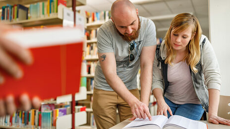 Photo of a female student and a male student in the library. He shows her something in an open book.__In the library, a student shows something to another student in an open book.