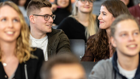 Close-up of several students in the rows of seats in a lecture hall. The focus is on a female student and a male student sitting next to each other and smiling at each other.<br>