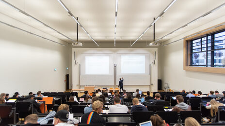 Von hinten Blick in einen vollen Hörsaal, vorne schreibt ein Lehrender etwas an die Tafel.