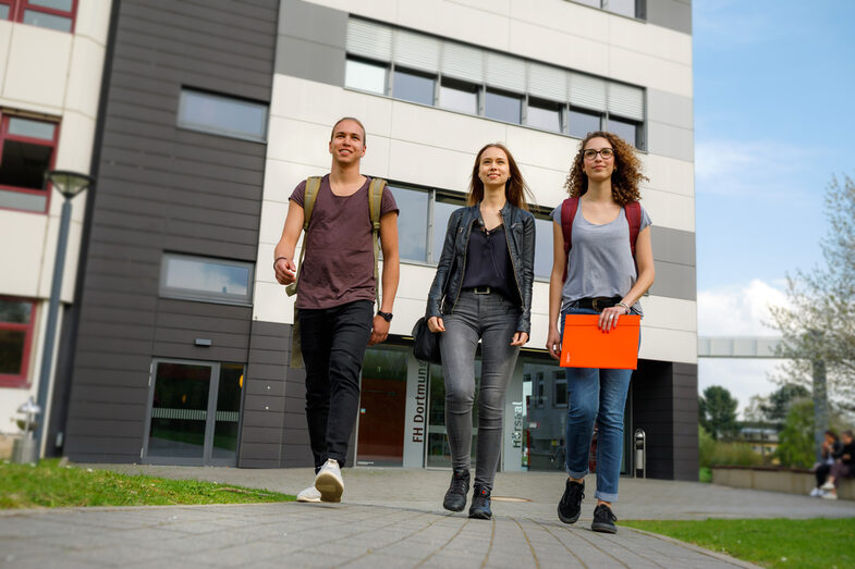 Photo of two female and one male student walking side by side on a path, in the background a building on Emil-Figge-Straße and other students __Two students walk side by side on a path, in the background a building on Emil-Figge-Straße and other students.