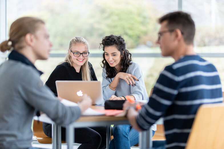 Foto von vier Studierenden an einem Gruppentisch. Im Fokus zwei Studentinnen, die nebeneinander sitzen und auf einen Laptop schauen. Zwei weitere Studenten sind in ein Gespräch vertieft.