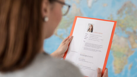 Photo over the shoulder of a woman. She is holding a folder with application documents or a CV in her hands. In the background a world map on the wall. __ Woman holds portfolio with application documents or a résumé in her hands. In the background a world map on the wall.