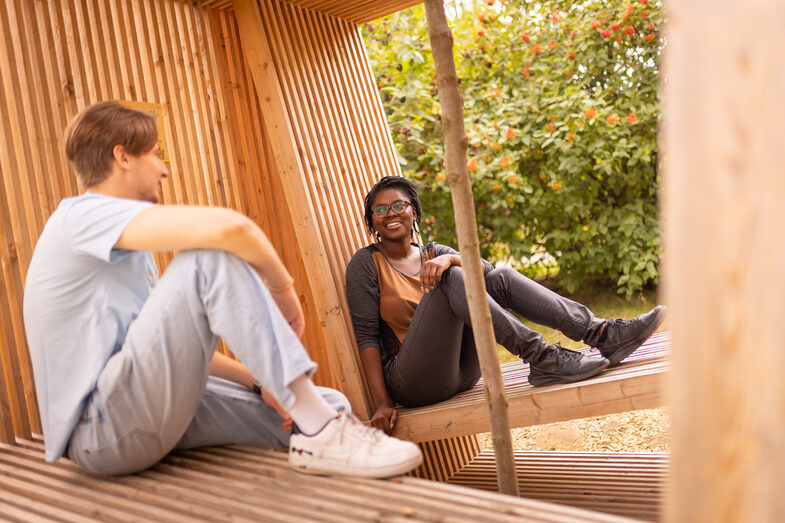 Photo of two students sitting in a wooden structure and chatting casually.