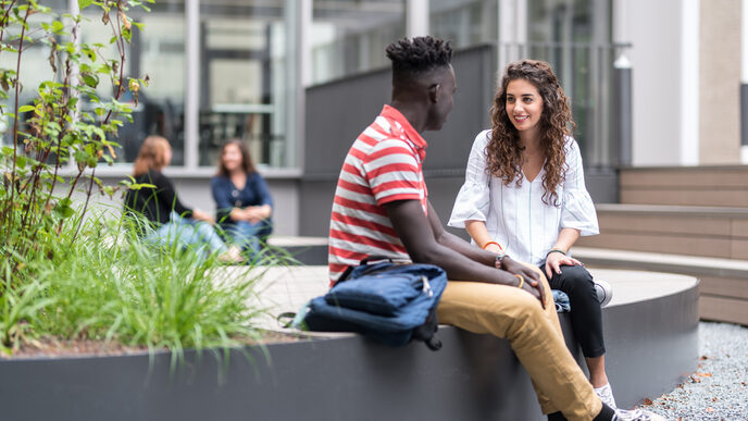 Photo of two students sitting and talking in the courtyard. In the background two other people sitting and a building. __ Young woman and young man are sitting in the inner courtyard and talking, in the background two other people and a building.