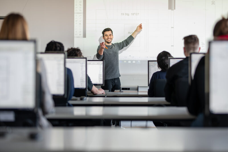Photo of a lecturer in the computer room. He is showing something on a graphic projected onto the wall. With his other hand, he smiles at a student as if he is calling that person. __ <br>An assistant professor is standing in the front of the computer room and is showing something on a graphic that has been projected onto the wall. With the other hand he shows a student smile as if he were calling that person.