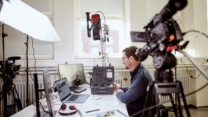 Photo of a lecturer sits at his desk, surrounded by various camera equipment as well as a laptop and monitors, in order to give an online lecture. __ A prof sits at his desk, surrounded by various camera equipment as well as a laptop and monitors, in order to give an online lecture.