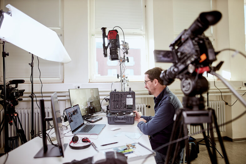 Photo of a lecturer sits at his desk, surrounded by various camera equipment as well as a laptop and monitors, in order to give an online lecture. __ A prof sits at his desk, surrounded by various camera equipment as well as a laptop and monitors, in order to give an online lecture.