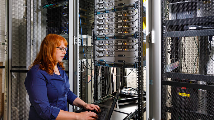 Photo of a trainee stands in front of a large cabinet with servers and operates a laptop. __ <br>A trainee stands in front of a large cabinet with servers and operates a laptop.
