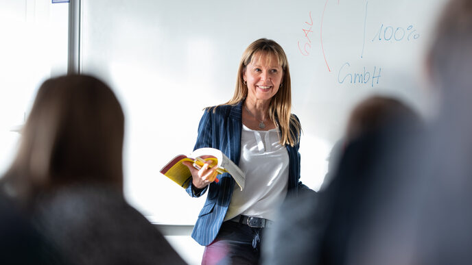 A professor stands smiling at the front of the room in front of a white board with notes. She holds a book in her hand and looks at her students sitting in the room. Only a few of the students are vaguely recognizable from behind. __ <br>A female professor stands in front of the room smiling in front of a white board with notes. She holds a book in her hand and looks at her students who are sitting in the room. Only a few of the students are out of focus from behind.