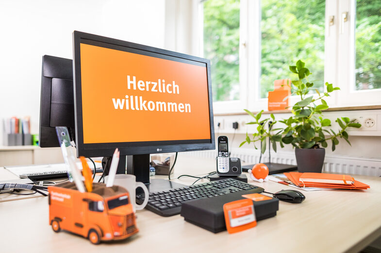 Photo of a desk with various orange-colored objects on it, e.g. a notepad, a name tag and a pen holder in the shape of the Citroën HY. On a PC screen, <br> "Welcome" is displayed. __A desk with various orange objects on it, e.g. a pad, a nameplate and a pen holder in the shape of the Citroën HY. "Welcome" is displayed on a PC screen.