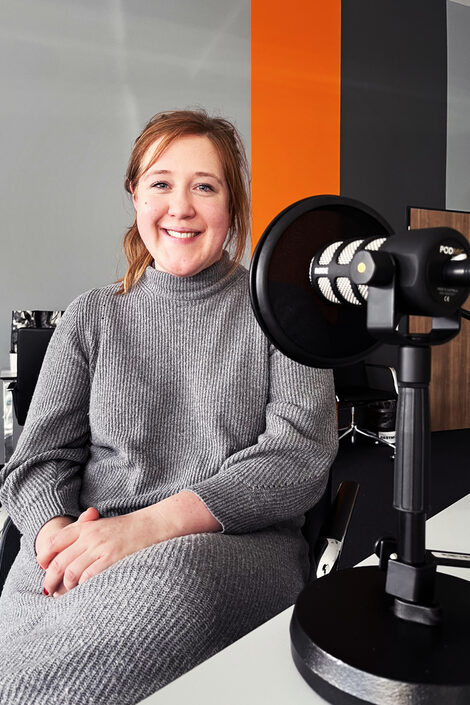 A person in a gray dress is sitting at a table with a microphone on it. The person looks into the camera with a smile.