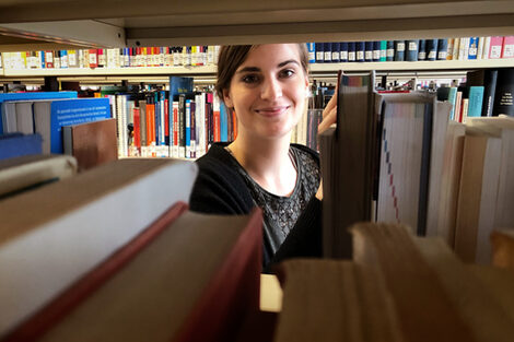 A woman looks through a bookshelf into the camera.
