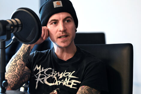 A man in a black T-shirt with a black cap and tattooed arms and hands is sitting at a table with a microphone on it. He looks past the camera on the right and appears to be in conversation with someone who is not in the picture.