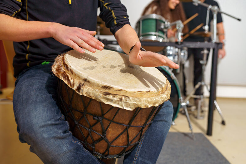 Photo of a man sitting and playing a djembe drum, his head is not in the picture. Two people are playing the drums in the background.