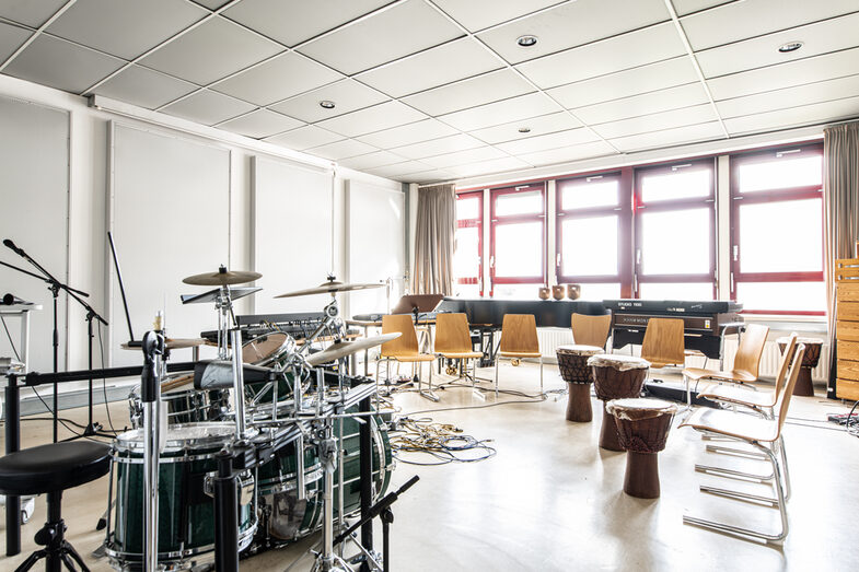 Photo of the audio lab. On the left is a drum kit and microphone stand. On the right are chairs and three djembe drums, at the back of the room is a grand piano.