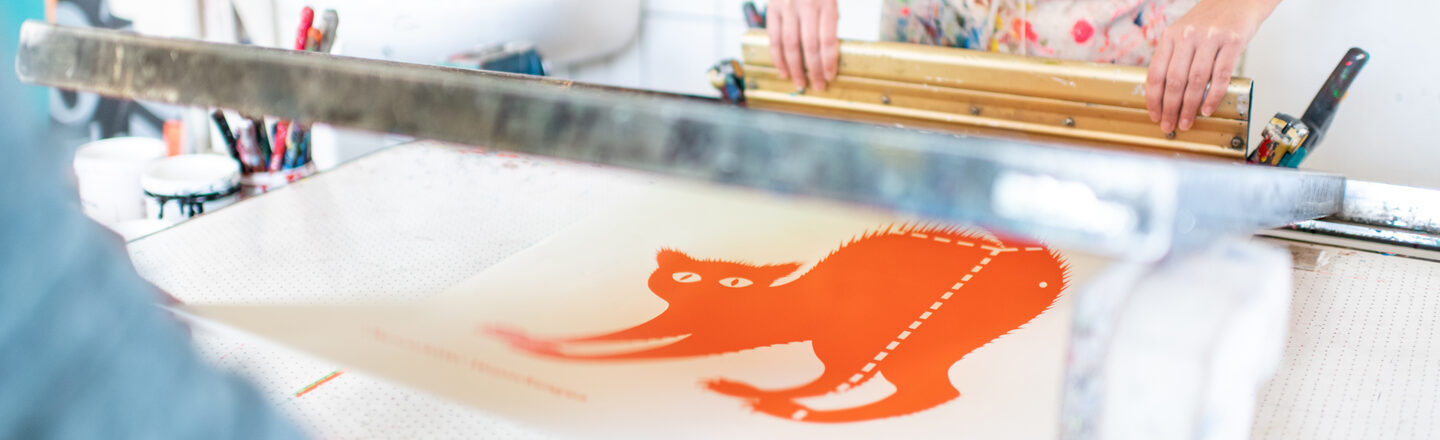 Photo of two people working together to create a screen print of an orange cat. Only the upper body and arms of the people can be seen.
