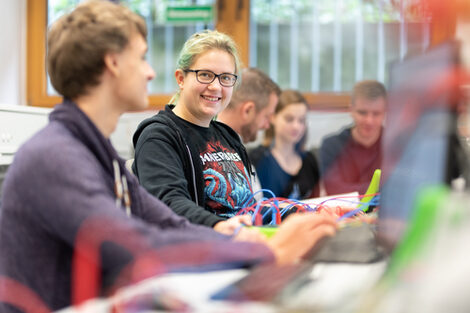 Photo of several students sitting next to each other at a row of tables with various electronic test setups. One student is in focus and smiling at the camera.