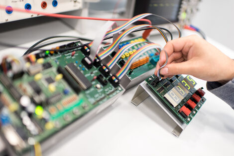 Photo of a male hand inserting a cable into an electrotechnical assembly with circuit boards.<br>On the front assembly are numbers on a field.