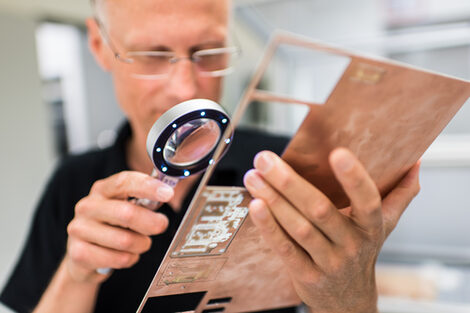 Photo of an employee of the Electronics and Automation Laboratory inspecting a milled circuit board with the aid of a magnifying glass.