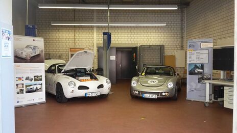 Photo of two cars in a workshop. The hood of one of the cars is open. There are displays with technical information about the cars in the workshop.