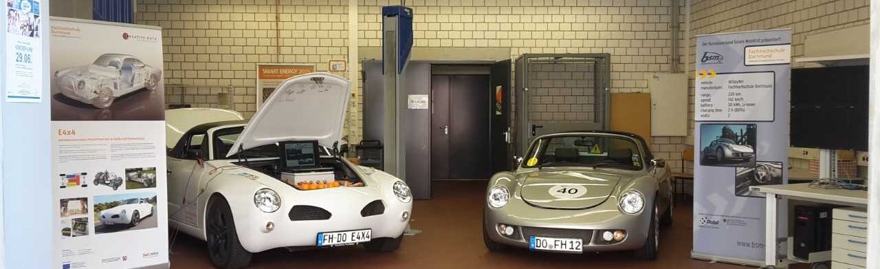 Photo of two cars in a workshop. The hood of one of the cars is open. There are displays with technical information about the cars in the workshop.