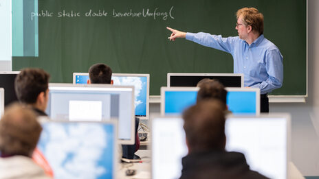 Photo of a professor showing something on the blackboard in the computer room. In front of him are students at computers.