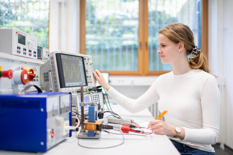 A photo of an experimental setup in which a coil on a laminated iron core is fed by a variable transformer via a sliding series resistor. A woman is sitting in front of the experimental setup and operating an oscilloscope with her right hand. She is taking notes with her left hand.