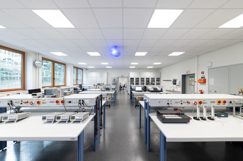 A photo of a laboratory room with many tables on which various devices are placed.<br>A projector hangs from the ceiling. On the side of the room you can see a window front.