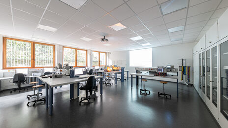 A photo of a laboratory room taken from the corner. There are experimental setups on the tables. In the background, the projector is beaming something onto the wall.