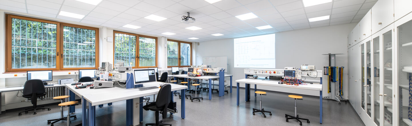 A photo of laboratory room A-113.1 taken from the corner. There are experimental setups on the three double tables. Four PC workstations can be seen on the left under the window and there are cabinets on the right wall, some with glass panes. In the background, the projector is projecting light onto the wall.