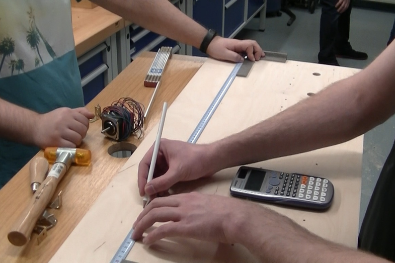 Photo of students building a floppy organ. One of them uses a ruler to draw a pencil line on a wooden panel. Only the arms of the people can be seen.