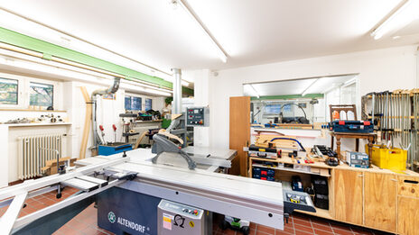 Room shot of part of the wood workshop with large circular table saw and other equipment in the room.