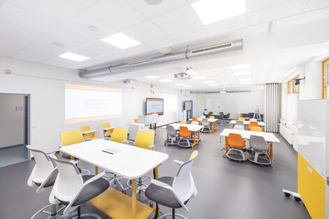 Image of the InnoLab, a modular and divisible room with various work areas. In the foreground is a high table with chairs, on the sides several digital whiteboards, a lectern and a projector surface as well as group tables. At the back there is a couch and a meeting table as well as walls to divide the room.