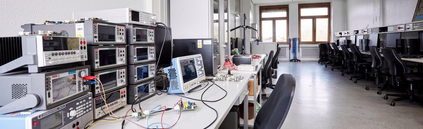 The picture shows an optics laboratory. You can see several workstations on the right and left. On the right-hand side, each workstation has a PC and on the left-hand side there are mainly other technical devices from electrical engineering, such as oscilloscopes.