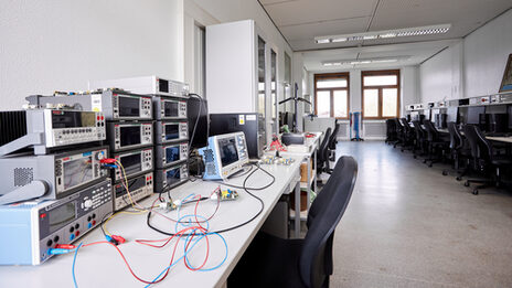 The picture shows an optics laboratory. You can see several workstations on the right and left. On the right-hand side, each workstation has a PC and on the left-hand side there are mainly other technical devices from electrical engineering, such as oscilloscopes.
