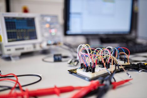 The focus in this picture is on a breadboard with several colorful cables attached to it. In the background you can see a switched-on oscilloscope and a switched-on PC screen.