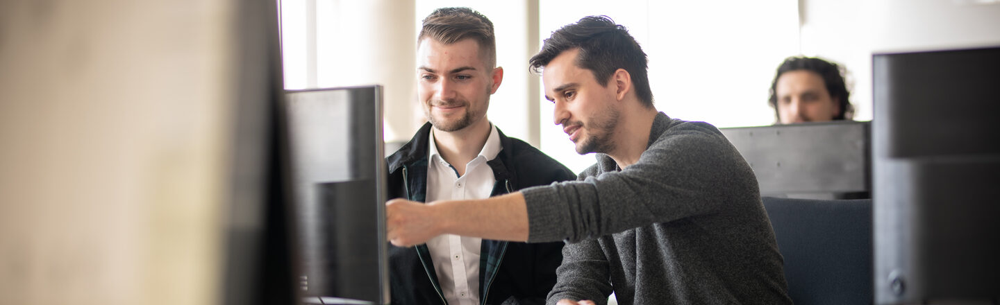 Photo of a lecturer next to a student at a PC. The lecturer is showing the student something on the screen. In the background is another student at a computer.