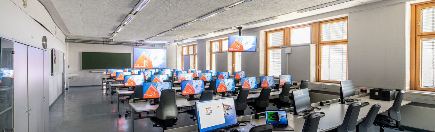 Photo of the computer room. There is a screen at the front of the wall. The last row of the room (at the front of the picture) are laptop workstations.