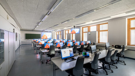 Photo of the computer room. There is a screen at the front of the wall. The last row of the room (at the front of the picture) are laptop workstations.