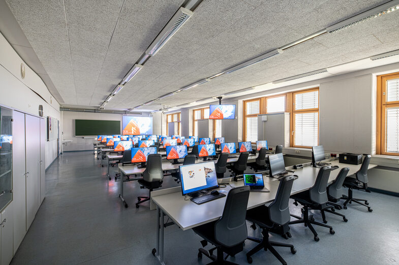 Photo of the computer room. There is a screen at the front of the wall. The last row of the room (at the front of the picture) are laptop workstations.