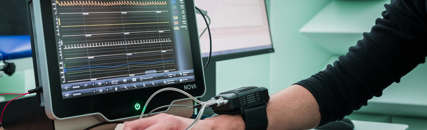 Photo of a hand with a connected measuring device on the table. Behind it, measurements are displayed or evaluated on a monitor.