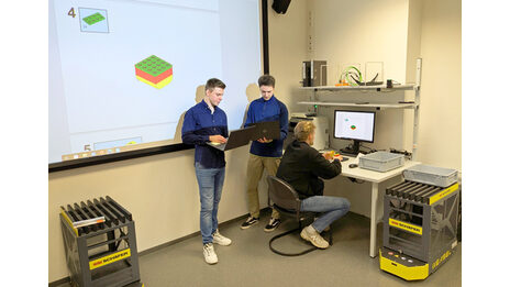 Zwei Personen stehen mit Laptops vor einer Leinwand. Eine dritte Person sitzt vor einem PC an einem Arbeitsplatz.