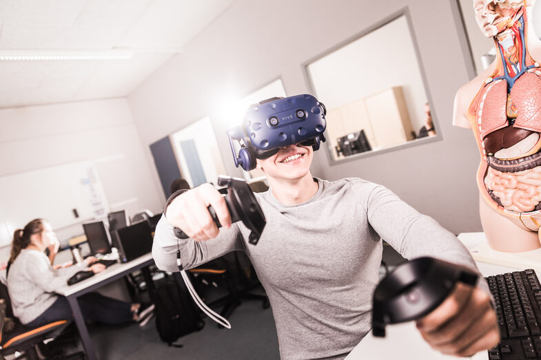 Photo of a student has VR glasses on and controller in his hands, is sitting at a computer workstation. __A student has VR glasses on and controller in his hands, is sitting at a computer workstation.